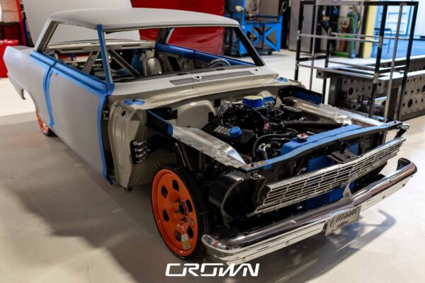 1964 Chevrolet Nova in primer with hood open, orange wheels, front three-quarter view at Crown Customs