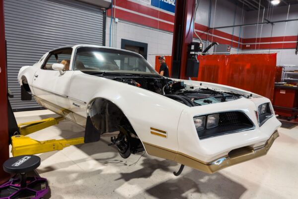 The white 1978 Pontiac Firebird Formula with gold lower accents raised on a lift at Crown Customs with its front wheels removed