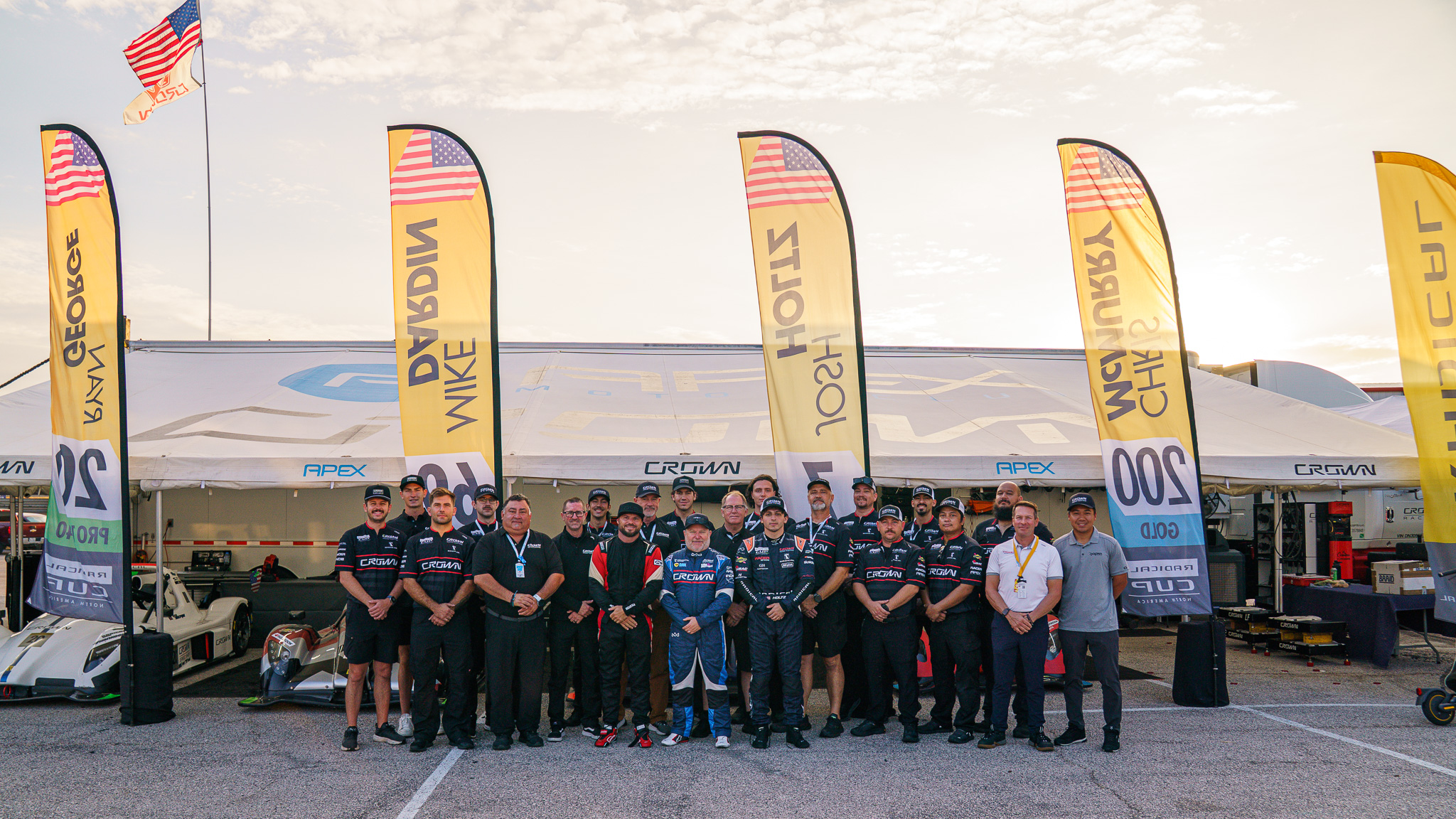 The Crown Racing crew and drivers in front of the Crown paddock tent at Circuit of The Americas, under driver banners for Mike Dardin, Josh Holtz and Chris McMurry