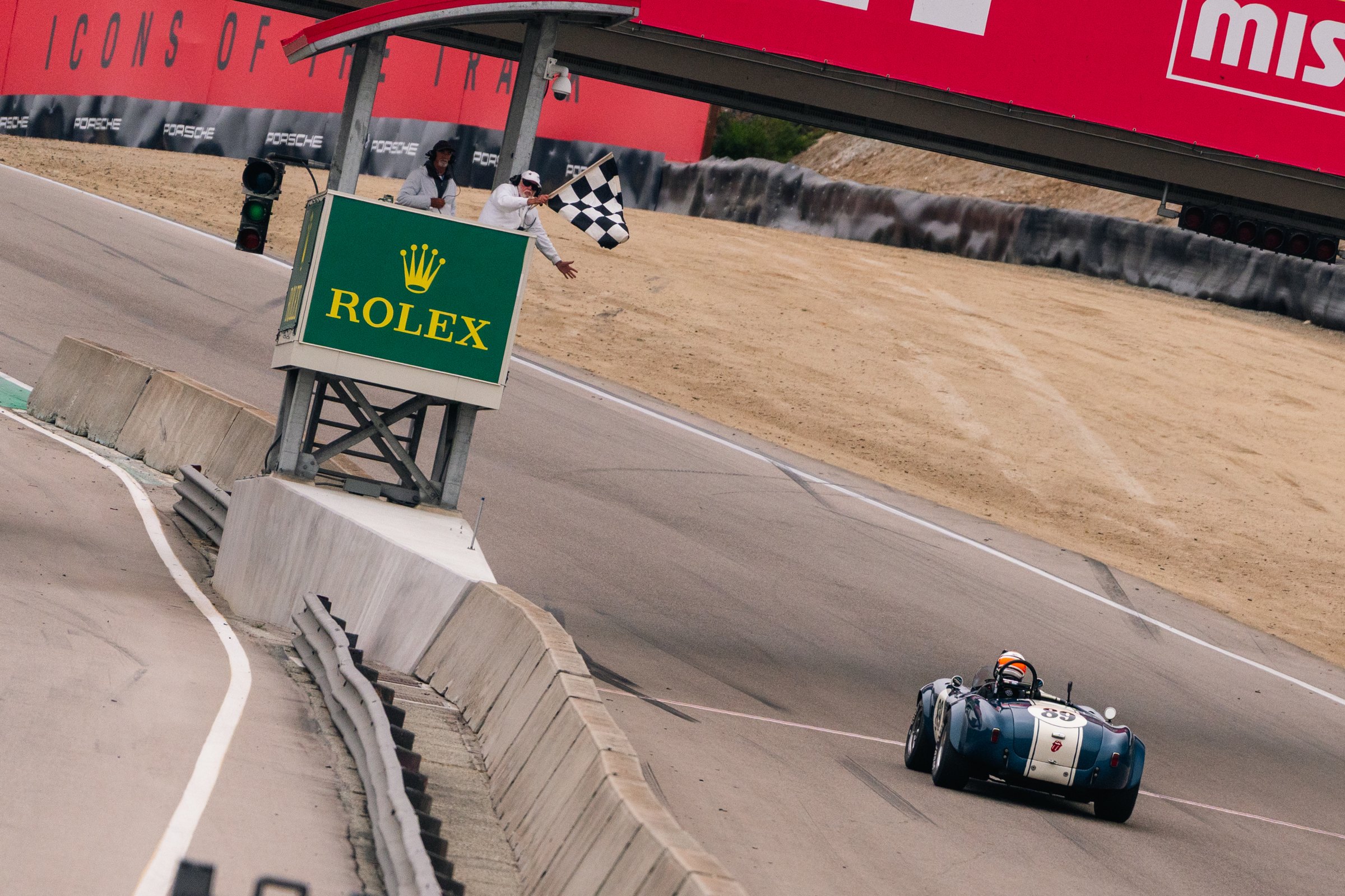 The blue number 89 1964 Shelby Cobra 289 takes the checkered flag beneath the Rolex flag stand at WeatherTech Raceway Laguna Seca