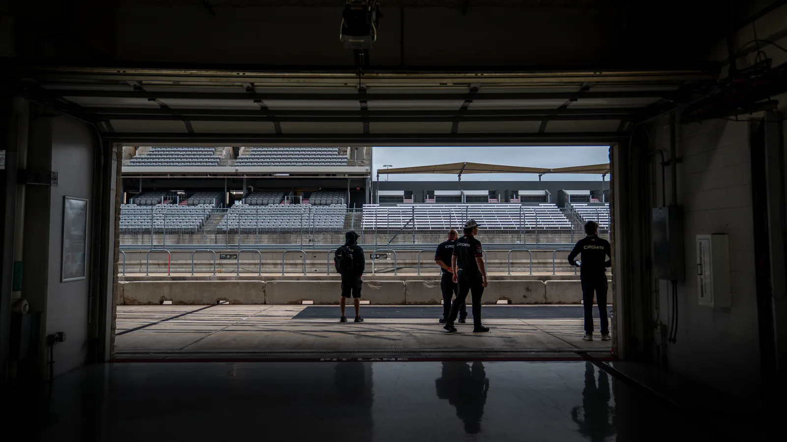 Crown Racing crew silhouetted in an open garage at Circuit of The Americas, looking out at the pit lane and grandstand before an IMSA session