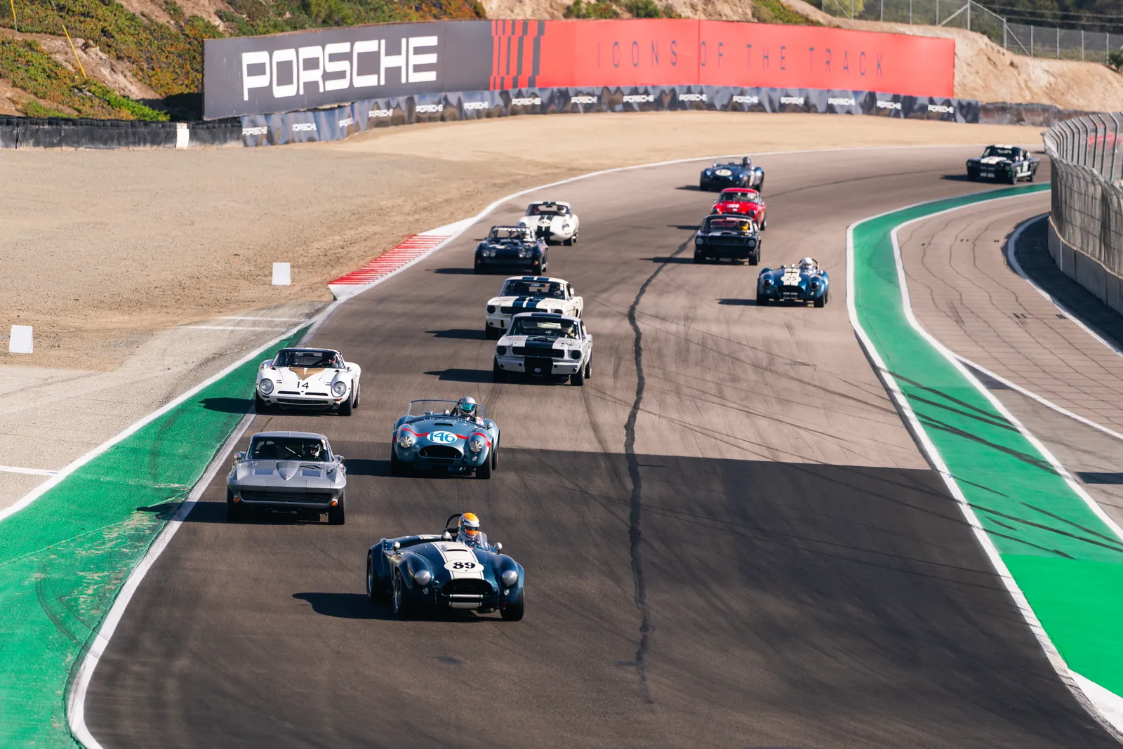 Field of historic race cars led by a Shelby Cobra, streaming under the Porsche bridge at Laguna Seca