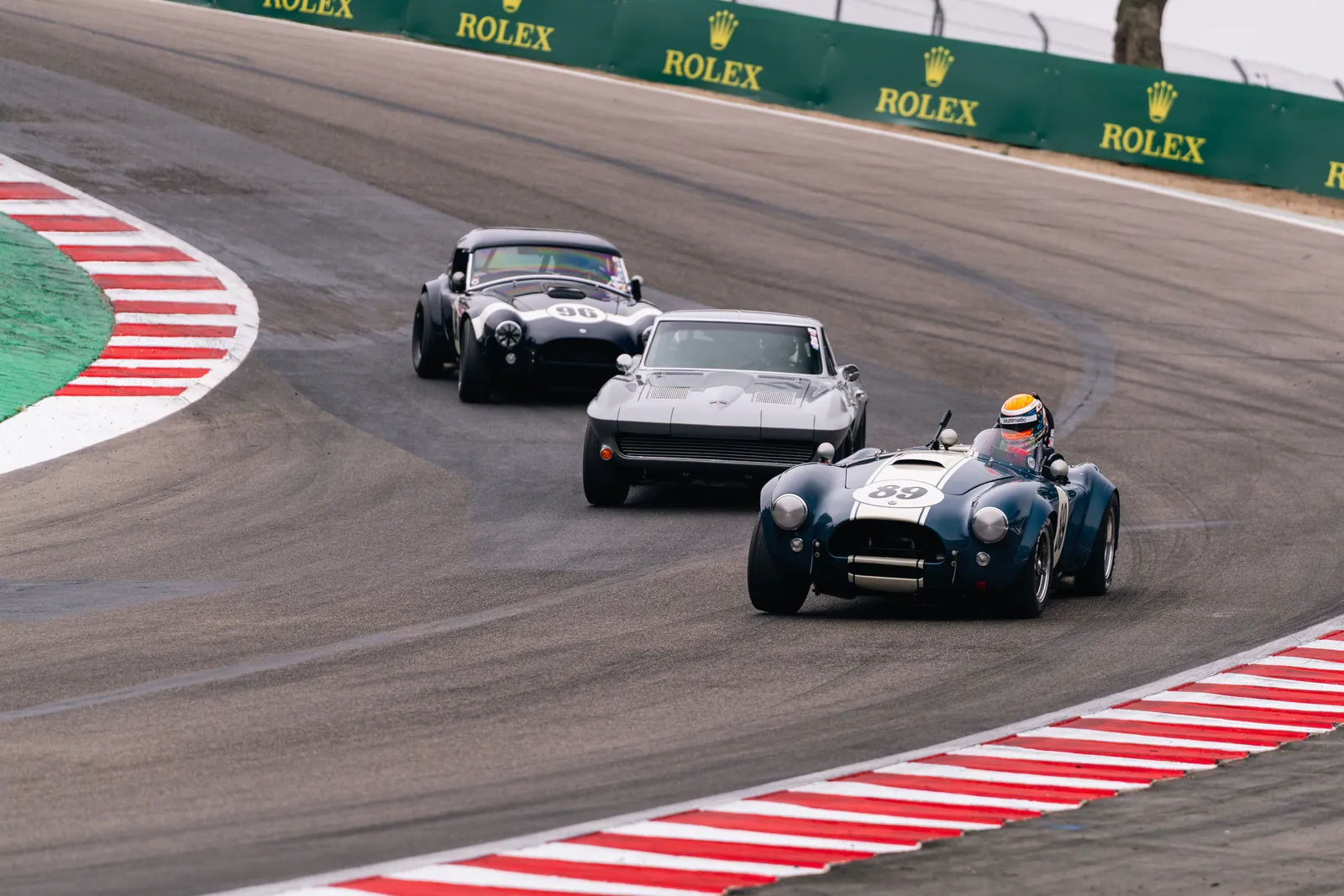 Blue No. 89 Shelby Cobra historic race car leading through a corner at Laguna Seca during the 2026 Rolex Monterey Motorsports Reunion, Rolex banners along the wall